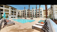 This outdoor amenity space showcases a thoughtfully designed resort-style courtyard anchored by a generously sized swimming pool with crystal-clear blue water. The pool deck is finished in warm-toned stone pavers and is lined with modern lounge chairs accented in turquoise cushions, creating a vibrant, inviting atmosphere. A separate spa pool is tucked to one side, adorned with a bold, colorful geometric tile mural that serves as a striking decorative focal point. Tall, swaying palm trees rise throughout the courtyard, adding a tropical character and providing natural shade across the space. The surrounding multi-story building features a Mediterranean-influenced facade with cream-toned stucco, arched entryways, private balconies, and large windows that look out directly over the pool area. Thoughtfully landscaped planter beds with tropical foliage, including banana plants and ornamental trees, line the perimeter and soften the transition between interior and exterior spaces. Floor-to-ceiling glass walls along the ground-level interior reflect the pool and courtyard, enhancing the sense of openness and connection to the outdoors. The overall ambiance is bright, airy, and resort-like, with an emphasis on outdoor living under abundant natural sunlight.