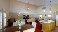 This single-floor space is designed with both comfort and functionality in mind, offering a seamlessly integrated layout that brings together multiple activity areas under one roof. The main living and dining area is anchored by soaring vaulted ceilings, which lend an airy, open feel to the room. An elegant chandelier hangs overhead, complementing the warm, neutral-toned walls and rich red carpeting that defines the space. The dining area features a dark wood round table surrounded by cushioned wicker chairs, accented by a decorative floral centerpiece. Adjacent to the dining space, the living area is furnished with plush cream-colored sofas and a large wooden entertainment center complete with a television, creating an inviting setting for relaxation and socializing. To one side, a kitchenette with a light-toned countertop and bar-height seating provides a convenient area for refreshments. A mirrored fitness room is also visible, outfitted with exercise equipment including a treadmill, offering residents a dedicated space for physical activity. The overall ambiance is warm, classic, and well-appointed, reflecting a thoughtfully designed communal environment suited for a variety of everyday activities.