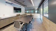 This conference room showcases a thoughtfully designed interior that combines industrial and contemporary aesthetics. The exposed concrete ceiling serves as a defining architectural element, complemented by black industrial pipe runs and linear pendant lighting that cast a clean, even illumination across the space. Patterned carpet tiles in neutral tones anchor the room and add a subtle layer of texture underfoot.

A large rectangular conference table in a warm taupe finish occupies the center of the room, surrounded by ergonomic mesh task chairs in light gray, providing comfortable seating for up to eight participants. Along one wall, built-in cabinetry in a light wood finish features a dark countertop, offering practical storage and surface space, with a wall-mounted flat-screen display positioned above for presentations and video conferencing.

A full glass wall with views into an adjacent workspace adds transparency and openness, while floor-to-ceiling windows along another wall invite abundant natural light and offer pleasant outdoor views of tree-lined surroundings. A solid wood-framed glass door with warm timber accents provides a welcoming entry point, reinforcing the room's balanced mix of raw and refined materials throughout.