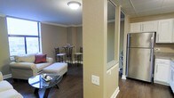 This single-floor space showcases a thoughtfully designed open-concept layout that seamlessly connects the living area and kitchen. Dark hardwood flooring runs throughout, providing a rich, warm foundation that contrasts elegantly with the neutral beige walls and crisp white trim and baseboards.

The living area is defined by its inviting arrangement, featuring a sofa with accent pillows, a cushioned ottoman, and a glass coffee table. A bistro-style high-top table with wrought iron chairs adds a casual dining element nearby, while a window along the wall allows natural light to filter into the space. Crown molding lines the ceiling throughout, lending a polished, finished look to the interior.

A textured structural column partially divides the living space from the kitchen area, which is visible to the right. The kitchen features stainless steel appliances, including a full-size refrigerator, white cabinetry, and what appears to be a granite or stone countertop. Mirrored panels near the kitchen passthrough add depth and visually expand the space.

Overall, the unit presents a cohesive, well-maintained interior with classic architectural detailing and functional, open living spaces.