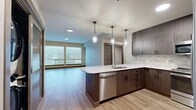 Located at 1940 W Riverstone Drive in the Riverstone neighborhood of Coeur d'Alene, Idaho, this single-floor residence offers a thoughtfully designed open-concept layout with contemporary finishes throughout. The kitchen serves as the heart of the home, featuring rich dark wood cabinetry, a textured tile backsplash, crisp white countertops, and a peninsula island with a built-in sink and stainless steel dishwasher. Elegant pendant lighting hangs above the island, adding a stylish focal point. Stainless steel appliances, including a range, microwave, and refrigerator, complete the well-appointed kitchen. Light-toned hardwood-style flooring flows through the main living and kitchen areas, reflecting natural light from large windows that provide views of the surrounding neighborhood. The living area is generously proportioned, offering an open and airy atmosphere. Bedrooms are finished with plush carpeting and feature windows that bring in natural light, with one bedroom offering sliding door access to an outdoor space. Sliding panel-style closet doors provide practical storage. Neutral wall tones and dark wood baseboards tie the design together, creating a cohesive, modern aesthetic suited for comfortable everyday living.