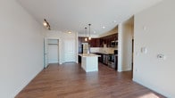 This single-floor residence offers a thoughtfully designed open-concept layout that seamlessly connects the kitchen and living areas. The kitchen is a standout feature, showcasing rich dark-espresso cabinetry, granite countertops, a tile backsplash, and a full suite of stainless steel appliances including a refrigerator, range, microwave, and dishwasher. A central island with a granite surface provides additional prep space and serves as a natural divider between the kitchen and the expansive living area. Warm hardwood-style flooring flows throughout the main living spaces, enhancing the inviting ambiance. The living area is bathed in natural light, courtesy of a large sliding glass door that opens onto a private balcony with outdoor views. The bedroom is generously sized, featuring plush carpeting, a ceiling fan with light fixture, and ample closet access. The bathroom is finished with a dual-sink vanity topped with a light countertop, dark wood cabinetry, a large framed mirror, and a combination tub and shower. Clean neutral wall tones and consistent finishes throughout contribute to a cohesive and modern aesthetic across the entire space.