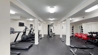 This gym offers a comprehensive fitness environment designed to accommodate a variety of workout routines. The space features white walls accented by crown molding and white structural columns, lending a clean, polished appearance throughout. Flush-mounted ceiling lights provide consistent, bright illumination across the entire floor area, ensuring excellent visibility during workouts.

The cardio section is well-appointed with multiple elliptical machines and a treadmill, ideal for endurance training. A wall-mounted television is positioned nearby, allowing users to stay entertained during cardio sessions. A centrally located cable weight machine serves as a focal point for strength training, offering versatility for a range of resistance exercises.

Along one wall, a dedicated free weights area features a tiered medicine ball rack stocked with a variety of weighted balls, complemented by additional strength training equipment. The flooring throughout is dark rubber, providing both durability and slip resistance. A wall clock and utility panels are integrated seamlessly into the space. Overall, the gym maintains a functional, organized layout with a tidy, professional ambiance.