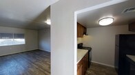 This single-floor space showcases a practical and straightforward layout, with an open connection between the kitchen and living area that promotes a sense of flow. The kitchen is equipped with warm medium-toned wood cabinetry, a black gas range with an overhead ventilation hood, and light-colored countertops that provide a clean contrast to the darker elements in the space. A textured white column serves as a partial divider between the kitchen and the adjacent living area, adding architectural definition without fully closing off the space. The living area is open and unobstructed, with neutral gray walls and dark hardwood-style flooring that extends throughout both areas, contributing to a cohesive and grounded aesthetic. Flush-mount ceiling light fixtures provide consistent overhead illumination in each area. Textured white ceilings and walls add subtle visual interest throughout. The overall ambiance is functional and understated, offering a clean, move-in-ready environment with a neutral palette that provides a versatile backdrop for a variety of interior styles.
