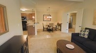 This single-story residence at Overbrook Apartment Homes offers an open-concept floor plan that seamlessly connects the kitchen, dining, and living areas, creating a cohesive and functional living environment. The kitchen is equipped with black appliances, including a range and built-in microwave, and features warm wood cabinetry, a tile backsplash, and a peninsula with an overhang that provides additional counter space and casual seating. The transition from the kitchen into the dining area is fluid, where a elegant multi-bulb pendant chandelier hangs above a dark wood dining table surrounded by upholstered chairs. A framed piece of artwork adorns the wall nearby, adding a decorative touch. The living area extends naturally from the dining space, anchored by plush carpeting in a neutral tone that runs throughout the main living zones, contributing to a warm and comfortable ambiance. Crown molding details along the ceiling add a refined architectural element. A doorway visible from the dining area suggests additional rooms beyond, including what appears to be a bathroom or utility space. Overall, the layout emphasizes openness, practicality, and understated elegance.