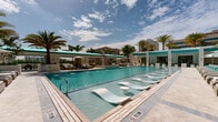Located in the heart of Coral Gables, Florida, this resort-style pool deck offers a luxurious outdoor retreat designed for relaxation and leisure. The centerpiece is a generously sized swimming pool with crystal-clear turquoise water, complete with built-in in-water lounge chairs that allow guests to cool off while soaking up the South Florida sun. Surrounding the pool, rows of cushioned chaise loungers are arranged on light-toned stone pavers, each paired with white umbrellas and accented with vibrant teal throw pillows that echo the color of the water. Lush tropical palms and manicured greenery border the deck, creating a sense of privacy and a verdant resort ambiance. A covered lounge pavilion with open archways and a translucent teal skylight ceiling provides a shaded gathering area furnished with seating and dining options, ideal for relaxing out of direct sunlight. The architectural design is clean and modern, characterized by crisp white walls, wide archways, and polished stone flooring that extends seamlessly between the indoor and outdoor areas. The entire space is framed by a high-rise residential building, reinforcing the upscale character of the community. The overall atmosphere is bright, airy, and reminiscent of a five-star hotel pool experience.