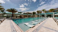 Located in the heart of Coral Gables, Florida, this resort-style pool deck offers a luxurious outdoor retreat designed for relaxation and leisure. The centerpiece is a generously sized swimming pool with crystal-clear turquoise water, complete with built-in in-water lounge chairs that allow guests to cool off while soaking up the South Florida sun. Surrounding the pool, rows of cushioned chaise loungers are arranged on light-toned stone pavers, each paired with white umbrellas and accented with vibrant teal throw pillows that echo the color of the water. Lush tropical palms and manicured greenery border the deck, creating a sense of privacy and a verdant resort ambiance. A covered lounge pavilion with open archways and a translucent teal skylight ceiling provides a shaded gathering area furnished with seating and dining options, ideal for relaxing out of direct sunlight. The architectural design is clean and modern, characterized by crisp white walls, wide archways, and polished stone flooring that extends seamlessly between the indoor and outdoor areas. The entire space is framed by a high-rise residential building, reinforcing the upscale character of the community. The overall atmosphere is bright, airy, and reminiscent of a five-star hotel pool experience.