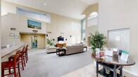 Located at 2900 W Highland St in Chandler, Arizona, this single-story clubhouse offers a well-appointed community gathering space designed for both relaxation and activity. The centerpiece of the clubhouse is a grand living area distinguished by soaring vaulted ceilings and an abundance of natural light streaming through multiple large windows and an arched transom window above. A teal accent wall adds a pop of color to the airy, open atmosphere. The living area is furnished with comfortable seating arrangements, a wall-mounted television, and a decorative fireplace with a tile surround, creating a warm and inviting ambiance. Southwestern-inspired décor, including wall art and lush indoor plants, adds character throughout the space. A reception-style table near the entry serves as an informational hub for residents. The kitchen area features warm wood cabinetry, granite-style countertops, and tile flooring, and is equipped with essential appliances including a coffee maker, microwave, and dishwasher — ideal for community events and gatherings. The dedicated exercise room offers a rubber-floored workout space stocked with a full dumbbell rack, kettlebells, a weight bench, cardio equipment, and a wall-mounted television, with direct access to the outdoors. Together, these spaces provide a functional and welcoming environment for the community.