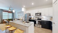 This single-floor residence offers a cohesive and modern living experience within a compact yet efficient footprint. The open-concept kitchen and living area serves as the heart of the home, where a striking two-tone kitchen commands attention with dark lower cabinets contrasting against crisp white upper cabinetry. Granite countertops extend across both the perimeter surfaces and a peninsula island with bar seating, complemented by a white subway tile backsplash and a full suite of stainless steel appliances. Pendant lighting above the island and recessed ceiling lights ensure the kitchen is well-lit throughout. Light-toned hardwood floors flow seamlessly into the living area, which is bright and airy, featuring large windows dressed with wood blinds and linen drapes that invite natural light. The bedroom is a calm, neutral retreat with plush carpet, large windows, a ceiling fan, and direct access to the bathroom and closet. The bathroom echoes the home's design language with granite countertops, white cabinetry, and clean finishes, including a soaking tub. Throughout the space, white walls and consistent finishes create a unified, modern aesthetic.