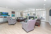 Located at 5460 Bentley Rd in West Bloomfield, Michigan, this single-floor property offers a thoughtfully designed, open layout that combines comfort and functionality. The centerpiece of the home is a generously sized living room showcasing a soaring vaulted ceiling with recessed lighting, warm light hardwood flooring, and crown molding throughout. A classic fireplace anchors the far wall, flanked by an impressive arrangement of floor-to-ceiling windows and French doors that flood the space with natural light and provide direct views of the surrounding greenery. The open-concept flow connects the living area to a dining space, which features a glass-top round table and additional workspace with wall-mounted framed artwork, creating a versatile area suited for both casual meals and focused tasks. A pass-through window into the kitchen — outfitted with warm wood cabinetry — maintains visual connectivity between rooms while preserving a defined sense of space. The living room is expansive enough to accommodate multiple seating arrangements, including two full-sized sofas and additional accent chairs, making it ideal for entertaining. Colorful artwork adorns the neutral-toned walls throughout, adding personality and vibrancy to the overall ambiance of this bright, well-appointed home.