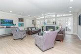 Located at 5460 Bentley Rd in West Bloomfield, Michigan, this single-floor property offers a thoughtfully designed, open layout that combines comfort and functionality. The centerpiece of the home is a generously sized living room showcasing a soaring vaulted ceiling with recessed lighting, warm light hardwood flooring, and crown molding throughout. A classic fireplace anchors the far wall, flanked by an impressive arrangement of floor-to-ceiling windows and French doors that flood the space with natural light and provide direct views of the surrounding greenery. The open-concept flow connects the living area to a dining space, which features a glass-top round table and additional workspace with wall-mounted framed artwork, creating a versatile area suited for both casual meals and focused tasks. A pass-through window into the kitchen — outfitted with warm wood cabinetry — maintains visual connectivity between rooms while preserving a defined sense of space. The living room is expansive enough to accommodate multiple seating arrangements, including two full-sized sofas and additional accent chairs, making it ideal for entertaining. Colorful artwork adorns the neutral-toned walls throughout, adding personality and vibrancy to the overall ambiance of this bright, well-appointed home.