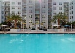 Located at 2263 SW 37 Ave in Miami, Florida, this amenity space serves as a well-equipped fitness center within a multi-story residential community. The fitness center is thoughtfully laid out across a single floor, offering a comprehensive selection of both cardio and strength-training equipment. Treadmills, a stair climber, and a stationary bike line one side of the room, catering to cardiovascular workouts, while a robust array of strength machines occupies the remaining floor space, providing options for a full-body training regimen. The interior features durable rubber flooring suited for high-traffic exercise use, recessed lighting, and clean white walls that create a bright, focused workout environment. Large windows and glass double doors allow natural light to filter in and provide views of the surrounding greenery and parking areas, enhancing the sense of openness. The fitness center connects directly to the community's outdoor amenities, most notably a expansive resort-style swimming pool framed by swaying palm trees and a paved deck with shaded umbrellas. The multi-story residential building serves as a striking backdrop to the pool area, reinforcing the cohesive, amenity-rich character of the community. Together, these facilities offer residents a convenient and complete wellness experience.