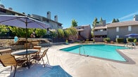 Located at 2380 Sierra Blvd in Sacramento, California, this outdoor amenity area serves as the central recreational hub of a multi-building residential community. The space is anchored by a generously sized swimming pool with clear, blue water, complemented by a separate hot tub/spa with a distinctive circular tiled design — ideal for year-round relaxation under Sacramento's abundant sunshine.

Surrounding the pool is a spacious concrete sun deck furnished with lounge chairs and umbrella-shaded patio tables and chairs, providing comfortable areas for sunbathing and outdoor socializing. The deck offers ample room for residents to spread out and enjoy the California climate.

The pool area is enclosed by a black iron perimeter fence with a gated entry, ensuring security and controlled access. An auxiliary structure adjacent to the pool area features a large window and panel siding, likely serving as a pool utility or storage facility.

Mature trees and ornamental shrubs are thoughtfully planted along the edges of the community, offering natural shade and greenery that soften the hardscape. The multi-story residential buildings visible in the background feature warm earth-tone exteriors with prominent chimneys and private balconies, framing the pool area and contributing to the community's cohesive architectural character. Pool rules signage is posted prominently, reflecting well-maintained community standards.