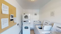 This laundry room is a practical, well-organized space designed to accommodate multiple users efficiently. The room features clean white walls and a light-colored ceiling with overhead lighting, creating a bright and functional environment. A stacked washer and dryer unit is positioned along one wall, maximizing vertical space, while additional freestanding top-load washers are arranged nearby, providing ample capacity for laundry needs. The flooring appears to be a neutral tile, offering durability and easy maintenance. A corkboard mounted on one wall serves as an informational hub, displaying posted notices and guidelines for users. An additional notice is affixed directly to the wall, ensuring important information is clearly visible. The layout is straightforward and efficient, with appliances arranged to allow comfortable access and movement throughout the space. The overall ambiance is clean, utilitarian, and well-maintained, reflecting a shared-use facility designed with practicality in mind.