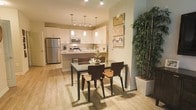This single-floor residence offers a thoughtfully designed open-concept layout that seamlessly connects the kitchen and dining areas, creating a cohesive and functional living environment. The kitchen is outfitted with white shaker-style cabinetry, stainless steel appliances including a full-size refrigerator and microwave, and a central island that provides additional workspace and casual seating. Pendant lighting hangs above the island, complementing a track lighting system that ensures the space is well-illuminated throughout. The dining area is defined by a sleek glass-top table paired with dark wood chairs, offering a contemporary yet inviting setting for meals and gatherings. Warm, light-toned wood-look flooring runs throughout, adding a sense of continuity and warmth to the space. Decorative touches, including framed wall art and a tall potted bamboo plant, bring personality and a natural element to the interior. The overall ambiance is modern and welcoming, with a neutral color palette of whites and warm beiges that enhances the sense of openness. The efficient single-floor plan makes the space easy to navigate, while the cohesive design ensures every area feels intentional and well-appointed.