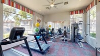 Located at 1320 South Val Vista Drive in Mesa, Arizona, this fitness center offers a well-organized, single-room workout space designed to accommodate a comprehensive range of exercise activities. The room is outfitted with a patterned, multi-toned carpet in shades of blue, red, and gray, adding a vibrant visual element underfoot.

Large windows adorned with colorful striped valances line the walls, flooding the space with abundant natural light and offering pleasant views of the surrounding landscaped grounds and community. A ceiling fan overhead provides additional air circulation, enhancing comfort during workouts.

The fitness center is equipped with multiple treadmills, a stationary recumbent bike, and a multi-station cable weight machine, catering to both cardio and strength training needs. A dedicated free weight area features a full dumbbell rack, while an assortment of exercise balls in various sizes adds versatility for flexibility and balance training. Wall-mounted mirrors expand the sense of space and allow users to monitor their form. A wall-mounted television is also present, providing entertainment during exercise sessions.

Colorful athletic artwork decorates the walls, contributing to an energetic and motivating atmosphere. The space is accessed through a glass-paneled door and is clearly marked as a Fitness Center, making it easy to locate within the property.