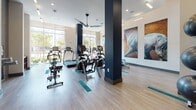 This single-floor fitness center is designed to support a comprehensive workout experience within a bright, open environment. The space is characterized by soaring ceilings and floor-to-ceiling windows that line the exterior walls, flooding the interior with natural light and offering views of surrounding greenery and outdoor landscaping. Light wood-tone flooring with teal accent strips runs throughout, providing a clean, modern aesthetic.

The facility is divided into distinct functional zones. The strength training area is equipped with cable machines, a multi-functional weight rack, adjustable benches, and a full dumbbell rack displayed in front of a large wall mirror, allowing users to monitor form. TRX suspension straps are mounted to a wall station, offering additional bodyweight training options. Nearby, bold navy accent columns provide architectural definition while delineating zones within the open floor plan.

The cardio section houses treadmills, ellipticals, and stationary bikes arranged to face the expansive windows, encouraging an energizing workout atmosphere. Ceiling fans circulate air throughout both areas. A separate glass-enclosed studio room, accessible via double glass doors, provides space for group fitness or focused training sessions. Large-scale abstract artwork adorns the walls, adding a curated, upscale ambiance to the overall space.