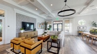 Located in Lake Worth, Florida, this single-story clubhouse at Garden Catalina Circle is a thoughtfully designed community gathering space that combines comfort with contemporary Florida style. The main lounge is an expansive, open-concept area anchored by a coffered ceiling with a dramatic circular pendant light. Wide-plank wood-look flooring, a stone-accented feature wall with a large mounted TV, and an arrangement of plush sectional seating and cognac leather chairs create a warm, inviting atmosphere. Abundant natural light pours in through large windows dressed in sheer white curtains, with glass doors opening directly to an outdoor patio area. Adjacent to the lounge, a dedicated dining area offers casual seating around natural wood tables, bathed in sunlight from multiple tall windows with views of lush Florida greenery. A built-in wet bar with a rich wood base, white countertop, and stylish wishbone bar stools provides a social focal point for entertaining. The game room is a standout feature, housing a full-size billiards table with a black felt surface beneath an artistic sculptural chandelier, complemented by a wall-mounted TV. A separate card room with a dedicated poker table, woven rattan chairs, and teal wingback seating opens to a private balcony, completing this versatile and elegantly appointed space.