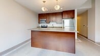 Located at 1249 Griswold in the heart of Detroit, Michigan, this two-bedroom, one-bathroom residence offers a thoughtfully designed single-floor layout. The kitchen serves as a central highlight, featuring rich dark wood cabinetry, light quartz-style countertops, stainless steel appliances including a refrigerator, electric range, and built-in microwave, and a peninsula with pendant lighting overhead — ideal for both cooking and casual dining. Light tile flooring with a distinctive mosaic accent border runs through the kitchen and bathroom, adding a refined decorative touch. The bathroom is sleek and modern, appointed with a glass-enclosed walk-in shower with tile surround, a dark wood vanity with an integrated sink and generous countertop space, and a large mirror. Both bedrooms are finished with neutral-toned carpet and soft gray walls, providing a calm and flexible canvas. Hallways connect the rooms efficiently, contributing to a practical flow throughout the space. The overall design palette — warm wood tones, neutral walls, and clean tile work — creates a cohesive, contemporary aesthetic well-suited to urban living in Detroit's Griswold corridor.