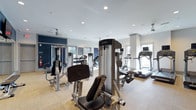 This fitness center is designed to support a comprehensive workout experience within a single, open-plan floor. The space is bright and airy, benefiting from multiple windows along the walls that allow natural light to fill the room, complemented by recessed ceiling lighting for consistent illumination throughout. The ceiling is finished in white with a clean, modern aesthetic, and a ceiling fan helps maintain comfortable airflow during workouts.

The layout accommodates a wide range of exercise equipment, including cable resistance machines, weight training stations, a dumbbell rack, treadmills, and additional cardio equipment such as ellipticals and steppers. Equipment is arranged to allow users to move freely between stations without congestion. A bold navy blue accent wall adds a strong visual focal point, contrasting with the otherwise neutral white walls and light-toned flooring.

Structural columns are integrated into the design, lending architectural character to the space. Large windows on multiple walls provide views to the outdoors, enhancing the overall sense of openness. The overall ambiance is clean, functional, and modern, creating an inviting environment for both strength training and cardiovascular exercise.