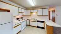 Located at 4330 Tenth Ave S in Fargo, North Dakota, this single-floor unit offers a practical and well-organized layout. The kitchen is equipped with white cabinetry trimmed in warm wood tones, providing generous storage across both upper and lower cabinets. It includes a electric range, dishwasher, refrigerator, and a double sink, all set against light tile flooring and a textured ceiling with overhead fluorescent lighting. The living area is open and airy, featuring plush carpeting and a sliding glass door flanked by vertical blinds that leads to an outdoor patio, allowing for natural light and exterior access. A wall-mounted air conditioning unit and baseboard heating ensure year-round comfort. The entry area transitions smoothly between the carpeted living space and the tiled kitchen zone, with two doors nearby providing access to additional rooms. The bedrooms are carpeted and feature warm-toned wood trim, with each room offering a closet fitted with a rod and shelf system for storage. The bathroom is clean and functional, featuring a single-sink vanity with wood cabinetry, tile flooring, and a full tub-shower combination. Throughout the unit, warm wood-toned doors and trim create a cohesive and inviting aesthetic.