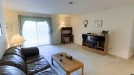 This single-floor space features a generously sized living room finished with wall-to-wall carpeting in a neutral tone, contributing to a comfortable and relaxed atmosphere. The room receives natural light through a large sliding glass door dressed with teal curtains, offering a bright and open feel. The layout comfortably accommodates a full seating arrangement, including a leather sofa and a wooden coffee table positioned at the center of the room. An entertainment center with shelving and storage drawers sits along one wall, providing practical media storage. A compact corner desk unit with an overhead hutch adds a functional workspace area within the living space. Decorative touches, including a wall clock and framed artwork, add character to the otherwise clean, neutral walls. Overhead ventilation is visible in the ceiling, suggesting a central HVAC system. The overall ambiance is understated and practical, with a straightforward layout that maximizes usable floor space throughout the room.
