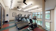 This single-floor fitness center is thoughtfully designed to support a comprehensive cardio workout experience. The space is outfitted with a full suite of Matrix brand equipment, including multiple treadmills, elliptical cross-trainers, and a recumbent stationary bike, all arranged to maximize usability and flow throughout the room. The flooring features a distinctive multi-tone pattern in shades of green, red, and cream, adding a vibrant energy to the environment. A row of large windows along one wall floods the space with natural light and offers pleasant outdoor views of landscaped grounds, creating an open and motivating atmosphere. The ceiling is fitted with a combination of linear LED lighting and ceiling fans, ensuring the room remains well-lit and comfortably ventilated during workouts. Decorative framed artwork adorns the walls, contributing a polished, inviting ambiance. Glass-paneled interior doors and partitions provide visual connectivity to adjacent areas while maintaining a sense of separation. The overall design strikes a balance between functional fitness utility and an aesthetically refined environment, making it a welcoming space for daily exercise.