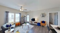 This single-floor residence offers a thoughtfully designed layout connecting its key living spaces through an open, flowing floor plan. The living room serves as the central gathering space, finished with dark hardwood-style flooring, neutral walls, and a ceiling fan overhead. Multiple windows allow natural light to fill the room, complemented by a door providing additional brightness. The space accommodates a comfortable seating arrangement alongside a media console and open shelving unit.

The kitchen is outfitted with white shaker-style cabinetry extending to the ceiling, stainless steel appliances including a refrigerator, range, and microwave, and a light-colored countertop island featuring an undermount double sink. Pendant lighting above the island adds a polished, modern touch. A stacked washer and dryer unit is also conveniently accessible from the kitchen area.

The bedroom is a well-proportioned retreat with dark hardwood-style flooring, a ceiling fan, and a window providing natural light and outdoor views. The bathroom features large-format marble-style wall tiles surrounding a full soaking tub and shower combination, white cabinetry for storage, and consistent neutral finishes throughout.