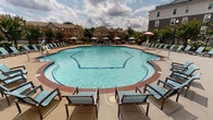 Located at 8825 Peregrine Heights Rd in Manassas, Virginia, this outdoor amenity space centers around a generously sized freeform swimming pool with a distinctive decorative tile border and clearly marked depth indicators. The pool's curvilinear design creates an inviting, resort-style atmosphere, complemented by a wide surrounding deck finished in neutral concrete with brick accent trim. Shallow entry points with stainless steel handrails provide convenient access throughout the pool perimeter.

The deck is thoughtfully appointed with an extensive collection of chaise lounges and armchairs in light blue, positioned to take full advantage of the open sky and sun exposure. Multiple patio umbrella stations in blue, teal, and red offer shaded seating areas with accompanying tables, ideal for relaxing or socializing. Decorative planters with seasonal flowers add color and greenery throughout the space.

The pool area is enclosed by a black iron fence, providing both security and a clear boundary while maintaining open sightlines to the surrounding neighborhood of multi-story residential buildings and mature trees. Decorative lampposts are positioned throughout the deck for evening illumination. The adjacent clubhouse building, constructed with a brick and dark siding exterior and featuring large windows, overlooks the pool and provides a handsome architectural backdrop to this well-appointed outdoor community amenity.