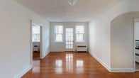 This single-floor residence offers a thoughtfully arranged layout that balances open living spaces with clearly defined functional areas. Throughout the home, warm-toned hardwood flooring adds richness and continuity, flowing seamlessly from room to room.

The main living area is bright and welcoming, featuring multiple windows that flood the space with natural light and offer views of the surrounding neighborhood. A door with glass paneling further enhances the airy feel, while a wall-mounted radiator provides efficient heating. The clean white walls create a neutral, versatile backdrop throughout.

The bedroom is a generously sized room with three windows along one wall, allowing ample natural light and views of greenery outside. A textured ceiling adds subtle architectural character, and a radiator ensures year-round comfort. The room also includes closet access and connects to additional spaces via an arched interior doorway, a charming architectural detail that adds classic character.

The kitchen is compact and efficiently designed, featuring tile flooring, rich dark wood upper cabinetry, a gas range, and a full-size refrigerator. A separate doorway leads to additional utility space, keeping the layout practical and organized.

Overall, the residence offers a cohesive and functional living environment, with classic architectural touches, well-maintained finishes, and plentiful natural light throughout.