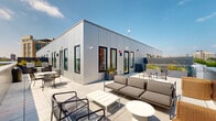 This rooftop amenity level offers a thoughtfully designed communal retreat that seamlessly blends indoor comfort with outdoor living. The interior lounge is bathed in natural light, courtesy of large windows dressed in sheer white curtains that frame sweeping city views. The space features a patterned tile floor with a botanical motif, smooth white walls, and clean-lined furnishings — including plush sofas, leather lounge chairs with slim metal frames, and accent stools — all arranged to encourage relaxed gathering. A large round mirror and circular wall sconces add refined decorative touches, while lush potted plants throughout infuse the space with vibrant greenery. A woven pendant light anchors the dining area, which is furnished with a solid wood table and modern chairs. The kitchenette is a bright, welcoming nook highlighted by white subway tile, open shelving lined with potted herbs and plants, globe pendant lights, and a clean white island with bar seating. Beyond the interior, expansive outdoor terraces wrap around the structure, offering multiple seating arrangements — from casual lounge sets to intimate bistro tables — all set against an open sky and panoramic urban skyline, making this an exceptional space for both relaxation and socializing.