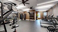 This fitness center is a thoughtfully designed workout facility that balances functionality with aesthetic appeal. The space is finished with warm, vertically slatted wood-paneled walls that lend a modern, upscale character to the room, complemented by a striking decorative wallpaper featuring an intricate botanical pattern on a contrasting accent wall.

The floor is covered in durable dark rubber flooring, well-suited for high-intensity use, while overhead, large ceiling fans and recessed linear LED lighting ensure the space remains comfortable and well-lit throughout. A wall-mounted mini-split air conditioning unit provides additional climate control.

The cardio zone is equipped with multiple TRUE-brand treadmills and elliptical machines, all featuring integrated display screens, positioned along the windowed wall to allow natural light and outdoor views during workouts. Stationary bikes are also available for cycling-based training.

The strength training area offers a multi-functional cable machine, a weight bench, and a freestanding rack stocked with medicine balls, kettlebells, and TRX suspension trainers. Full-length mirrors along one wall expand the visual depth of the space and allow users to monitor their form. The combination of premium equipment, natural light, and refined design details makes this fitness center a standout amenity.