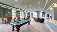 Located at 700 Citation Lane in Cherry Hill, NJ, this single-story community clubhouse offers a thoughtfully designed gathering space built for leisure and entertainment. The main hall is a generously proportioned open area finished with elegant gray walls, white wainscoting, tray ceilings with recessed lighting, and a patterned carpet throughout. Large multi-pane windows line the perimeter, flooding the interior with natural light and providing views of the surrounding grounds. A full-size billiards table anchors the game area, illuminated by classic green pendant lights, while multiple seating arrangements — including a large curved sectional sofa and several round tables with chairs — provide flexible areas for lounging and card or board games. A dedicated bar area features an expansive granite countertop, dark cabinetry with glass-front uppers, a wall-mounted flat-screen television, and pendant lighting overhead. An adjoining café-style kitchen is equipped with warm wood cabinetry, a coffee station, and additional counter space for serving. Wall sconces, framed artwork, and a decorative upright piano add refined touches throughout, creating an inviting and versatile community amenity space.