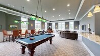 Located at 700 Citation Lane in Cherry Hill, NJ, this single-story community clubhouse offers a thoughtfully designed gathering space built for leisure and entertainment. The main hall is a generously proportioned open area finished with elegant gray walls, white wainscoting, tray ceilings with recessed lighting, and a patterned carpet throughout. Large multi-pane windows line the perimeter, flooding the interior with natural light and providing views of the surrounding grounds. A full-size billiards table anchors the game area, illuminated by classic green pendant lights, while multiple seating arrangements — including a large curved sectional sofa and several round tables with chairs — provide flexible areas for lounging and card or board games. A dedicated bar area features an expansive granite countertop, dark cabinetry with glass-front uppers, a wall-mounted flat-screen television, and pendant lighting overhead. An adjoining café-style kitchen is equipped with warm wood cabinetry, a coffee station, and additional counter space for serving. Wall sconces, framed artwork, and a decorative upright piano add refined touches throughout, creating an inviting and versatile community amenity space.