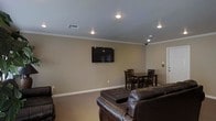 This single-floor space offers a comfortable and functional layout designed for both relaxation and casual dining. The main living area is anchored by a set of dark leather sofas and a tufted ottoman, arranged to face a wall-mounted flat-screen television — creating an inviting lounge atmosphere ideal for unwinding or gathering with others. Recessed lighting is installed throughout the ceiling, providing consistent and even illumination across the room. Crown molding runs along the upper perimeter of the walls, adding a clean, polished architectural detail that elevates the overall finish of the space. The walls are painted in a warm neutral tone that complements the earthy palette of the furnishings and the beige carpeted flooring. Adjacent to the lounge area, a modest dining setup features a dark wood table accompanied by matching wooden chairs, offering a dedicated space for meals or casual conversation. A visible HVAC vent in the ceiling ensures climate control throughout. The space is straightforward in design yet thoughtfully appointed, blending practicality with a welcoming ambiance suited to everyday use.