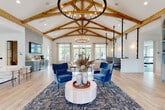 This expansive single-story communal space is defined by its dramatic vaulted ceilings adorned with exposed natural wood truss beams, lending a warm, modern-farmhouse character throughout. A large wrought-iron chandelier anchors the main gathering area, where light hardwood floors extend seamlessly across the open layout. Abundant natural light filters through multiple sets of French doors and large windows, offering views of the surrounding greenery and outdoor areas.

The main lounge features multiple seating arrangements, including plush blue velvet accent chairs grouped around a round pedestal coffee table, neutral sofas, and a leather bench, creating versatile spaces for both socializing and relaxation. A striking built-in shelving unit finished in muted blue with arched display niches serves as a focal point along one wall, while a living green wall framed in black steel grid adds a bold, organic accent nearby.

A dedicated dining and kitchenette area showcases sage green cabinetry, a dramatic vertical grey tile backsplash, and a geometric pendant light, with direct access to the outdoors. Suspended booth-style seating with slim black poles provides additional private workspaces. The overall ambiance is polished yet inviting, blending contemporary and farmhouse-inspired design elements throughout.