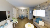 This single-floor residence offers an open-concept layout that integrates the living and dining areas into one cohesive, functional space. Hardwood-style flooring runs throughout, lending warmth and visual continuity across the entire unit. Neutral gray-toned walls provide a versatile backdrop, complemented by pops of teal, green, and yellow introduced through furnishings and décor.

The living area is comfortably proportioned, featuring a gray sofa accented with teal throw pillows and a round white coffee table. Adjacent to it, a white lattice chair with a green cushion adds a casual, welcoming touch. The dining area is defined by a round white table surrounded by blue chairs, centered beneath a modern drum pendant light. Wall-mounted planters above add a decorative, organic element.

A partial wall with floating shelves separates the dining space from a secondary area, which appears to transition into a hallway leading to additional rooms. A front entry door is visible, reinforcing the practical flow of the layout. The space is well-lit through a combination of artificial lighting and ambient brightness, creating an inviting and lively atmosphere overall.