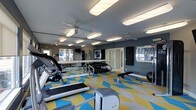 This fitness center is designed to accommodate a wide range of workout needs within a single, open-plan space. The room is finished with a bold geometric-patterned floor in blue, yellow, and grey, adding a dynamic, energetic atmosphere to the environment. The walls are painted in a deep charcoal grey, complemented by large mirrors that line one side of the room, enhancing the sense of space and allowing users to monitor their form during exercise.

Overhead, multiple flush-mounted rectangular light fixtures provide bright, even illumination throughout, while ceiling fans promote air circulation and comfort during intense workouts. The space is equipped with cardio machines, including treadmills and a stationary bike, positioned toward the front of the room. A dedicated free weights area features a full dumbbell rack with an accompanying bench, suitable for strength training routines. A stability ball and additional exercise equipment round out the offerings.

Windows along the perimeter allow natural light to filter in, contributing to an open, welcoming feel. The overall layout is functional and well-organized, catering to both cardio and strength training.