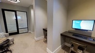 This single-floor space presents a thoughtfully designed layout with a neutral, professional aesthetic. The workspace area features two built-in dark-wood desks positioned against structural columns, each accompanied by metal chairs and equipped with monitors, making them well-suited for individual or shared computer use. The columns, finished in a clean white base with smooth gray walls above, add architectural definition while naturally segmenting the workspace into semi-private stations. The flooring throughout is a textured, light-toned carpet that contributes a quiet, focused atmosphere. Crisp white baseboards contrast neatly against the warm gray walls, lending the space a clean, polished appearance. Recessed lighting provides consistent, even illumination across the area. An adjacent room, visible through a black-framed glass door, reveals cabinetry in a light finish and lighter flooring, suggesting a utility or storage area that complements the primary workspace. The overall ambiance is calm and functional, with a cohesive color palette and understated design details that support productivity without distraction.
