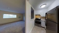 This single-floor space presents a straightforward and functional layout, with distinct areas for living and cooking visible from a central corridor. The kitchen is equipped with stainless steel appliances, including a gas range with hood ventilation and a refrigerator, complemented by white cabinetry that provides ample storage. The light-toned tile flooring in the kitchen adds a clean, practical finish. A pass-through window connects the kitchen to the adjacent living area, enhancing the sense of openness between the two spaces. The living area features wall-to-wall carpeting in a neutral tone, contributing to a comfortable and understated atmosphere. A window unit air conditioner and a baseboard heater are present, ensuring year-round climate control. Natural light filters into the living space through a window on the exterior wall. The walls throughout are finished in a soft, neutral palette, keeping the overall aesthetic clean and simple. The flush-mounted ceiling light provides straightforward illumination. The space is well-suited for everyday living, with a practical design that prioritizes utility and ease of use.