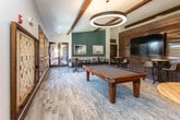 Located at 2071 Powers Ferry Rd SE in Marietta, Georgia, this single-floor community amenity space offers a well-appointed gathering area designed for both socializing and relaxation. The interior showcases a warm, contemporary aesthetic defined by exposed dark wood ceiling beams, light hardwood-style flooring, and a striking shiplap wood accent wall that anchors a large flat-screen television — an ideal centerpiece for communal viewing. A full-size pool table serves as a focal point of the recreational zone, complemented by bar-height tables and modern upholstered stools arranged throughout for casual conversation and dining. The lounge area features plush gray sofas and armchairs, creating comfortable seating clusters. A standout oversized wall-mounted Scrabble board adds a playful, interactive element to the space. Vaulted ceilings and an abundance of large, dark-framed windows flood the interior with natural light and offer views of the surrounding mature tree canopy and outdoor deck. Teal accent walls and curated artwork contribute to a vibrant yet cohesive design palette. A glass-door entry leads to an outdoor deck, further extending the usable gathering space and enhancing the connection to the natural surroundings.