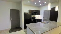 This single-floor space showcases a thoughtfully designed kitchen as its focal point. The kitchen features a striking contrast between dark espresso-toned cabinetry and crisp white walls, creating a bold yet clean aesthetic. Upper and lower cabinets provide generous storage, while a suite of black appliances — including a full-size refrigerator, electric range, and built-in microwave — offers a cohesive, contemporary look.

A standout feature is the granite-topped kitchen island or peninsula, finished in a light speckled stone that balances the darker elements of the space. The integrated sink adds functionality to this surface. Recessed lighting throughout the ceiling ensures the kitchen is well-lit, enhancing the polished finish of the countertops and cabinetry hardware.

The flooring transitions between tile in the kitchen area and wood-look planks in the surrounding space, adding visual interest and practical durability. Neutral white walls keep the space feeling open and bright. Two interior doors are visible — one appearing to be an entry door and another leading to an additional room — suggesting a practical, well-connected layout throughout this single-floor residence.