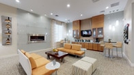 This well-appointed single-floor communal lounge space offers a sophisticated and welcoming environment designed for both relaxation and social interaction. The open layout is anchored by a striking wall-mounted linear fireplace set against a polished glass-panel surround, creating a refined focal point in the main seating area. Warm amber and neutral tones are carried throughout via plush upholstered sofas, accent chairs, and patterned ottomans, all arranged atop a textured, neutral-toned carpet that adds depth and warmth underfoot.

Recessed lighting and pendant fixtures illuminate the space evenly, while large windows dressed with sheer layered curtains invite abundant natural light. A dedicated refreshment station lines one wall, featuring warm wood cabinetry, glass-front upper cabinets, a wall-mounted flat-screen television, and a counter equipped with multiple beverage appliances. High-top bistro tables and bar-height seating provide casual dining options alongside the refreshment area.

An adjoining room, visible through a glass-door entryway and framed by a decorative chandelier, offers additional seating and display space, suggesting a flexible use area or reception zone. Curated artwork and decorative accents are placed throughout, contributing to the polished, contemporary aesthetic of the overall space.