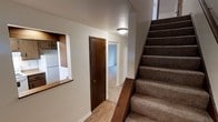 This three-floor residence offers a well-organized layout designed for practical, everyday living. The main level sets the tone with a combination of hardwood-style flooring in the entryway and hallway areas, providing a warm and inviting introduction to the home. A carpeted staircase with a classic wooden banister connects the floors, offering both functionality and a traditional aesthetic touch.

A notable architectural detail on the main level is a pass-through window that opens between the hallway and an adjacent room — likely the kitchen — complete with visible cabinetry above, suggesting a convenient connection between living and dining or food preparation spaces. Dark-stained wooden doors throughout the main level add contrast against the neutral, light-toned walls, contributing to a clean and understated interior palette.

The hallway leads to additional rooms carpeted in a neutral tone, hinting at comfortable, private living spaces beyond. Flush-mount ceiling lighting provides consistent illumination throughout the common areas. The overall ambiance is straightforward and functional, with a neutral color scheme that offers a versatile canvas. Spread across three full floors, this residence provides ample space for a variety of living arrangements and uses.