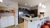 The Bristol is a single-floor, one-bedroom, one-bathroom residence designed with functionality and style in mind. The kitchen serves as a central highlight, showcasing dark cabinetry that contrasts sharply with light wood-style flooring and a granite-look countertop peninsula. Stainless steel appliances, including a full-size refrigerator, built-in microwave, and a gas range with a mosaic tile backsplash, give the kitchen a polished, contemporary feel. Recessed lighting illuminates the space efficiently, enhancing the clean aesthetic. The peninsula provides additional prep and casual dining space, opening the kitchen toward the living area for an inclusive, connected layout. Just off the kitchen, a dedicated laundry area houses a full-size washer and dryer, adding notable convenience. Upper cabinet space extends to the ceiling, maximizing storage throughout the kitchen. The overall design balances practicality with modern finishes, creating a cohesive and comfortable living environment well-suited for everyday living.