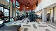 Located at 4015 Craft St in Charlotte, NC, this contemporary fitness center is designed to deliver a premium workout experience across a single, well-appointed floor. The main workout area immediately impresses with its dramatic double-height ceilings finished in a bold terracotta-red accent, complemented by large windows that flood the space with abundant natural light and views of the surrounding exterior. Stylish black pendant lights and ceiling fans hang overhead, lending an industrial-modern aesthetic throughout.

The patterned, geometric carpet tile flooring defines the expansive main floor, which is fully equipped with a wide range of strength-training machines, cable systems, free weights, and functional fitness equipment. Full-length mirrors line portions of the walls, enhancing the sense of openness and supporting form-focused training. Flat-screen TVs are integrated into the equipment areas for entertainment during workouts.

A dedicated cardio alcove features multiple treadmills positioned beside large windows with courtyard views, creating an airy and motivating environment. Dark cabinetry and built-in storage provide organized accessory space within the room.

The entry area welcomes visitors with clean white walls, curated large-format photographic artwork, and patterned flooring that carries through from the main floor. Glass-paneled entry doors with dark metal framing create a polished, transparent facade, offering a welcoming first impression to this well-equipped fitness facility.