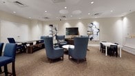 This single-floor community space is thoughtfully designed to accommodate both social gatherings and practical daily needs. The central lounge area serves as the heart of the space, featuring a carefully curated arrangement of high-back blue upholstered chairs grouped around a white coffee table, complemented by wooden benches and additional seating. Recessed lighting illuminates the neutral-toned walls, which are adorned with striking abstract artwork, while a wall-mounted flat-screen television anchors one end of the room. The carpeted flooring adds warmth and comfort throughout the lounge.

Adjacent to the social area, a fully equipped kitchen is outfitted with stainless steel appliances, including a refrigerator, electric range with overhead microwave, and a built-in microwave and oven combination. Crisp white cabinetry with dark hardware pairs with dark countertops, creating a clean, functional workspace. The kitchen features tile flooring and ample overhead lighting.

The facility also includes a dedicated hallway with dual water fountains positioned at varying heights for accessibility, alongside clearly marked restroom access. The overall layout, as seen in the aerial overview, reflects a practical and welcoming design suited for communal use.