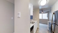 This single-floor, 2-bedroom, 2-bathroom residence at Spruce is designed with practicality and modern comfort in mind. The kitchen features white cabinetry, light stone-patterned countertops, and a stainless steel sink, complemented by a bold blue tile backsplash that adds a pop of color to the clean, neutral palette. Under-cabinet space accommodates a dishwasher, maximizing efficiency in the compact yet well-organized kitchen layout. Light wood-look flooring runs throughout, creating a cohesive and easy-to-maintain surface across the living spaces. An intercom panel is conveniently mounted near the kitchen entry, adding a layer of practical functionality. The adjacent living area is bright and airy, benefiting from a large window that draws in natural light and offers exterior views. A ceiling fan with an integrated light fixture ensures year-round comfort. The overall design aesthetic is clean and contemporary, with neutral wall tones and modern fixtures that provide a versatile backdrop for a variety of furnishing styles. The thoughtful layout and modern touches make this residence a comfortable and well-appointed home.