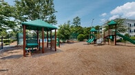 Located at 4632 Still Meadow Dr in Wilmington, NC, this outdoor community amenity space offers a well-rounded recreational environment designed for residents of all ages. The centerpiece of the area includes two tennis courts — one with a traditional green surface and another with a blue and green hard court finish — both enclosed by chain-link fencing and equipped with overhead lighting for extended play hours. A white bench sits courtside, providing a comfortable spot for spectators.

Adjacent to the courts is a generously sized playground area surfaced with protective wood chip mulch. The playground features a multi-level play structure with dual slides, climbing elements, and a covered gazebo-style structure with seating — ideal for younger children. A swing set arch and additional freestanding play equipment round out the area, which is enclosed by fencing for safety.

The grounds are thoughtfully landscaped with mature crape myrtles, ornamental grasses, flowering shrubs, and manicured hedgerows. Meandering gravel pathways wind through the greenery, offering a peaceful setting for leisurely strolls. The lush canopy of trees provides ample shade throughout the space, enhancing the overall ambiance. The natural surroundings and well-maintained grounds create an inviting, active outdoor environment for the community.