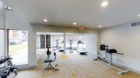 Located in Fort Worth, this fitness center offers a thoughtfully designed, single-floor workout space that balances functionality with a clean, modern aesthetic. The interior features neutral-toned walls and recessed lighting that work in tandem with large windows along the far wall, flooding the space with natural light and providing open views of the surrounding exterior.

The layout is spacious and well-organized, accommodating a variety of exercise equipment. The cardio zone is prominently featured and includes multiple treadmills and elliptical machines positioned near the windows, allowing users to exercise with an outward view. A dedicated strength and functional training area is equipped with a medicine ball rack, an adjustable workout bench, and cable or resistance machines for targeted strength work.

Flooring throughout the space consists of patterned carpet tiles with accent coloring, providing comfort underfoot while defining workout zones. A central support column subtly divides the room into distinct areas without disrupting the open feel of the space. Recessed ceiling lights and an illuminated exit sign contribute to a safe and well-lit environment. Overall, the fitness center presents a practical and inviting atmosphere suited for a range of workout routines.